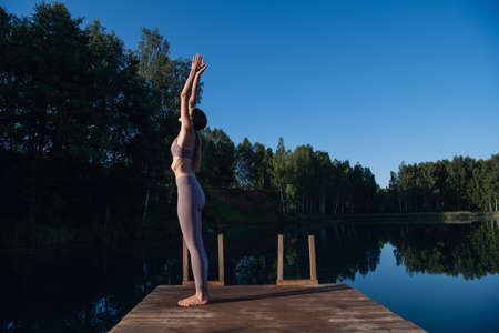 Young female practicing yoga against the sky on a sunrise. Pretty slim woman performs an exercise. Healthcare, meditaion, welness, mindfulness concept.の写真素材