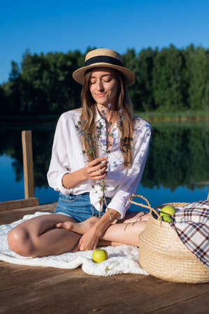 Portrait beautiful stylish romantic retro girl holding flower branch in hand while sitting at pier near the lake outdoors in nature.の写真素材