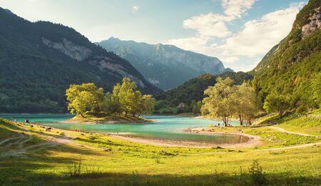 Lago di Tenno (Italy, Alps) - beautiful landscape with mountain lake (with island), beach and grassy meadow in foreground, mountains and blue sky with white clouds in background, Summer sunny dayの写真素材