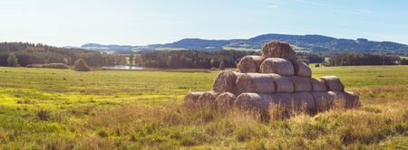 stack of straw bales on field of grass, landscape with meadow, pond, forest and hills in background, summer day with clear skyの写真素材