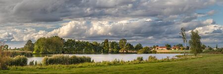 picturesque country landscape with pond, meadow and trees, in background village and beautiful white cloudsの写真素材