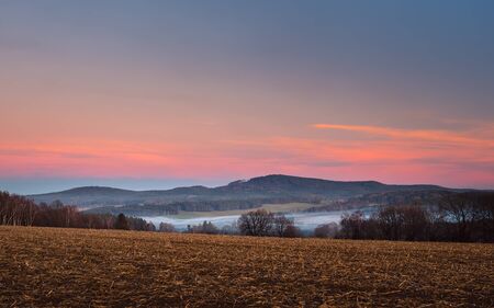 landscape at sunset, in foreground field and trees, in valley white fog, in background forest and...