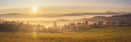 sunset in the mountains - hilly landscape with meadows and forests in a haze, into a yellow and orange-colored skyの写真素材