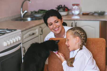 Mom and her little daughter playing with a cat in the kitchenの写真素材