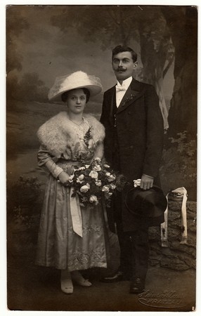 CHRASTAVA (KRATZAU), THE CZECHOSLOVAK  REPUBLIC - CIRCA 1920s: Vintage photo of newlyweds. Bride wears a furry shawl and  white wide- brimmed hat with netting. Groom wears posh clothing, white bow-tie, gloves and black top hat.  Black & white antique studのeditorial素材
