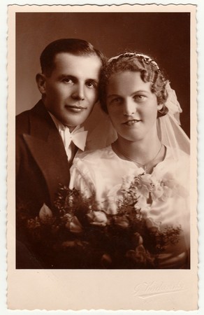 CHRASTAVA (KRATZAU), THE CZECHOSLOVAK  REPUBLIC - CIRCA 1920s: Vintage photo of newlyweds with wedding bouquet. Bride wears wedding veil headdress. Groom wears posh clothing, white bow-tie. Black & white antique studio portrait.のeditorial素材
