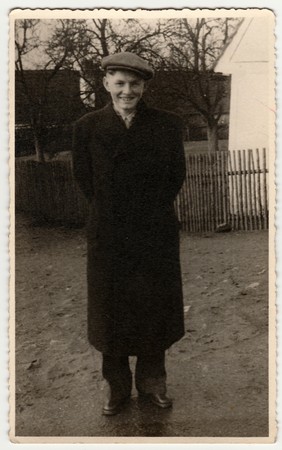 THE CZECHOSLOVAK SOCIALIST REPUBLIC - CIRCA 1950s: Vintage photo shows young man wears peaked cap, poses outdoors. Wooden fence and house is on background.  Black & white photography.のeditorial素材
