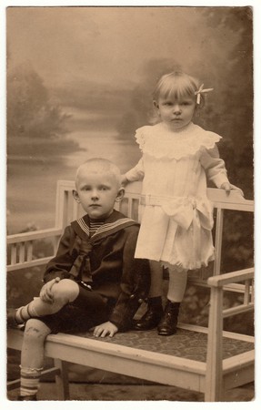 HRADEC KRALOVE, THE CZECHOSLOVAK REPUBLIC - AUGUST 11, 1930: Vintage photo shows two children (siblings). Boy wears sailor costum and girl wears white dress, laced collar and hair ribbon. They pose on historic white bench. Black & white studio photographyのeditorial素材
