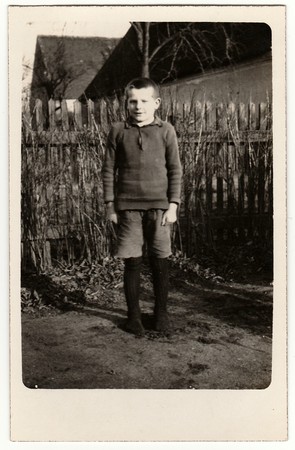 GERMANY - CIRCA 1940s: Vintage photo shows young boy (pupil, student) stands in front of wooden fence. Black & white antique photography.のeditorial素材