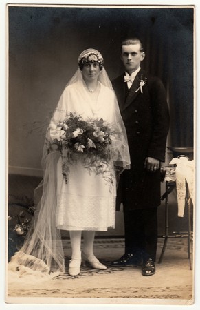 CHRASTAVA (KRATZAU), THE CZECHOSLOVAK  REPUBLIC - CIRCA 1930s: Vintage photo of newlyweds. Bride wears a long veil and holds wedding bouquet. Groom wears black suit and white bow tie. Black & white antique studio portrait.のeditorial素材