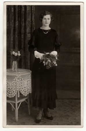 BERLIN, GERMANY - CIRCA 1940s: Vintage photo shows woman wears black dress and she holds bouquet in hands. Studio photography contains the small table and crocheted tablecloth.のeditorial素材