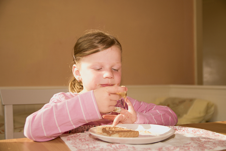 Kid girl playlful eats chocolate cream spread on bread. Chocolate sweet food snack. Happy girl has a snack in the kitchen. A cute small girl smiles. A small girl with chocolate cream stains on face. Childhood and fun concept.の写真素材
