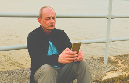 Midle aged man sitting on the shore of the lake. Solitude mature man using mobile phone on the bank....