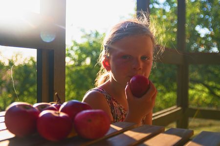 The little girl is sitting at a table on a verandah and eatting fresh apples. Apples on table. Dreamy and romantic image. Summer and happy childhood conceptの写真素材