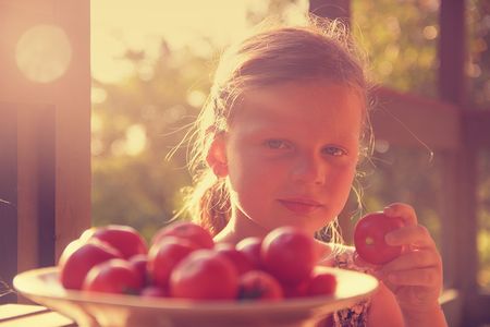 The little girl is sitting at a table on a verandah and eatting fresh tomatoes.の写真素材