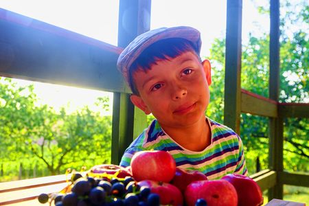 Little boy with fruit in the garden. Little smiling farmer boy wearing a flat cap and eating organic fruits, grapes, apples. The concept of harvest. Dreamy and romantic image. Autumn and happy childhood conceptの写真素材