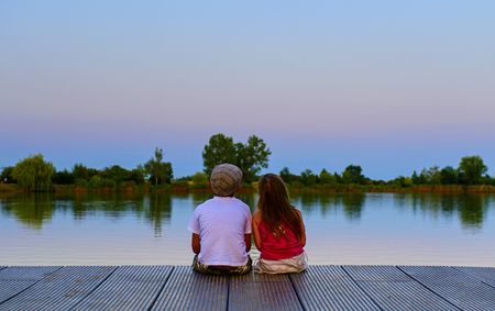 Boy with flat cap and little girl are sitting on pier. Boy and girl are looking on red sky. Love, friendship and childhood concept. Beautiful romantic sunset pictureの写真素材