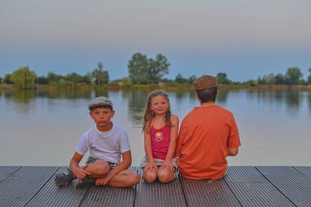 Children sitting on pier. Siblings. Three children of different age - teenager boy, elementary age boy and preschool girl sitting on a wooden pier. Summer and childhood concept. Children on bench at the lake. Golden hour during vacation evening.の写真素材