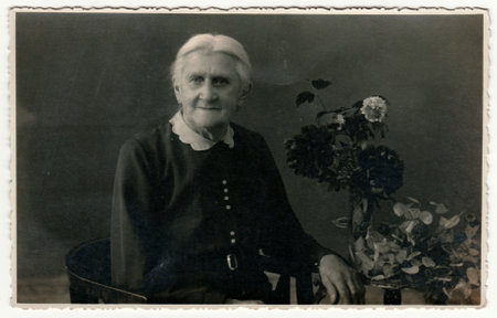 GERMANY - CIRCA 1930s: Vintage photo shows elderly woman sits on the period armchair in a photography studio. On the table are flowers. Retro black and white studio photography with sepia effect.のeditorial素材