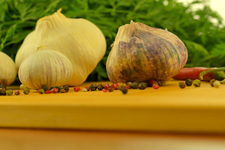 A bulb of garlic, red chili peppers, four-color pepper, and carrot tops on a wooden cutting board. The concept of cooking and vegetables in the home. Macro image, close-up. Garlic - Allium sativum is a species of bulbous flowering plants in the genus Allium. Its close relatives include the onion, shallot, leek, chives, Welsh onion, and Chinese onion. Garlic is native to central and south Asia, stretching from the Black Sea through the southern Caucasus, northeastern Iran, and the Hindu Kush, it also grows wild in parts of Mediterranean Europe. There are two subspecies and hundreds of varieties of garlic.の写真素材