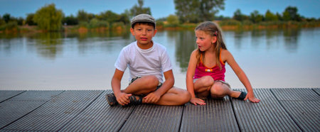A group of cute little children sit on a wooden pier by a lake. Small children looking into the future. Children sit on a dock. Siblings. Two children of different ages a school-age boy and a preschool-age girl sitting on a wooden deck. The concept of summer and kids. Golden hour during a vacation evening. The concept of summer, vacation, and carefree childhood.の写真素材