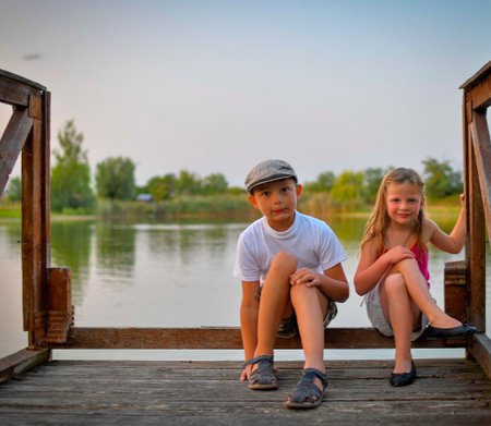 A little boy and girl sit on a wooden log on the shore of a lake. Small children sit on a pier at the edge of a pond. The concept of a carefree childhood and summer vacation.の写真素材