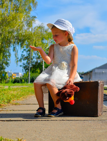 A lovely little girl in a white dress with a cuddly toy sits on an old suitcase. A sweet smiling girl on a train platform. The concept of summer holidays, travel, and a carefree childhood and familyの写真素材