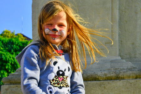A cheerful young girl with playful bunny face paint and a teddy bear hoodie enjoys a breezy day...