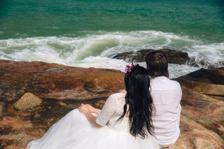 groom in white suit and the bride in a dress holding each others hands  standing on the background of the sea. beautiful landscape with big stones. concept wedding dayの写真素材