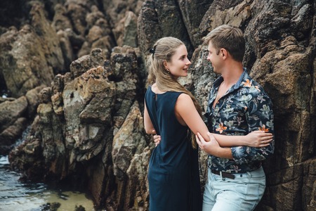 young couple in love in black shirt holding hands on the beach and looking at the sea. big stones background. Concept of familyの写真素材