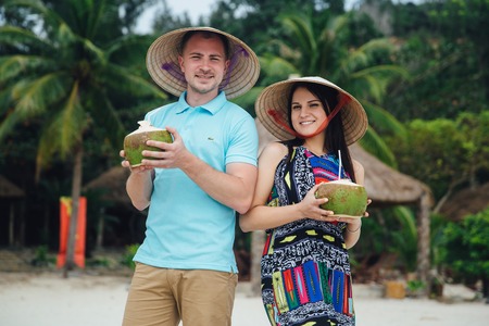 Portrait of young smiling beautiful couple in Vietnameme hats holding coconut in hands. beautiful beach with palm trees in the backgroundの写真素材