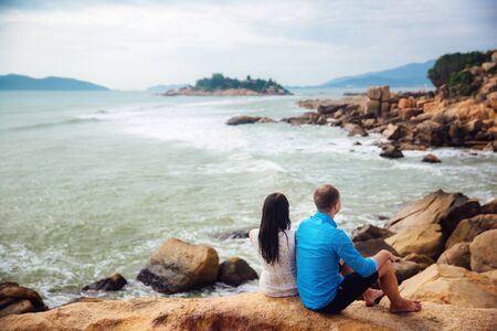 Young couple in love sitting at big stones back to back on a sea beach in summer. man wear the blue shirt and the girl in a white dress with a wreath in her hairの写真素材