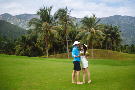 Young beautiful couple in love holding coconut in hands and walking at golf course in summer. man wear the blue shirt and the girl in a white dress and vietnamese hats. Concept of honeymoonの写真素材