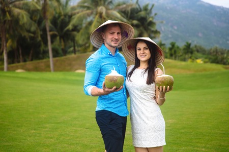 Young beautiful couple in love holding coconut in hands and walking at golf course in summer. man wear the blue shirt and the girl in a white dress and vietnamese hats. Concept of honeymoonの写真素材