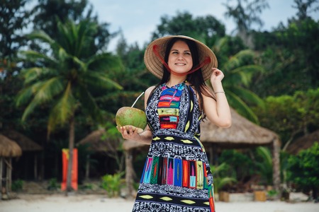 Portrait of young smiling beautiful brunette woman in a colored dress and Vietnameme hat holding coconut in hands. beautiful beach with palm trees in the backgroundの写真素材
