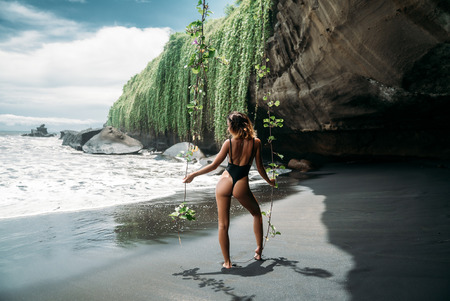 Rear view, a girl with a sexy body and an ass dressed in a black swimsuit resting on the beach with black volcanic sand. Beautiful young model with a sporty body and tanned skin posing on a sea beach with large waves.の写真素材