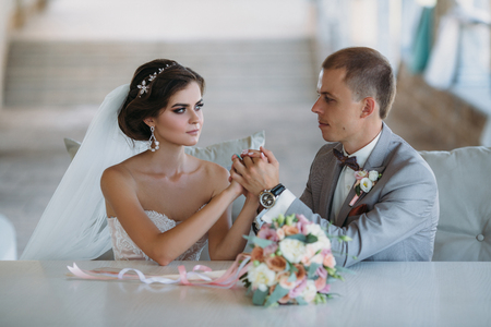 beautiful wedding couple hugging in park with green trees on background. Groom in a business gray suit, white shirt in a bow tie and a buttonhole. Bride in a luxury dress with a veil and a bouquet of flowers. Wedding couple holding each others hands.の写真素材