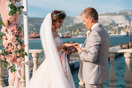 couple in wedding arch exchange rings with lake on background, the bride with long beautiful hairs and groom in black suit look at each other in wedding day. Concept of love and family