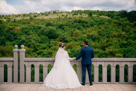 A girl in a lavish wedding dress is standing near a stone bridge and holding her husbands hand. The newlyweds admire the hills.の写真素材