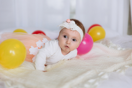 A newborn girl lies in a white bandage and dress on a bed with colored balls. The child himself holds his head and studies the world.の写真素材