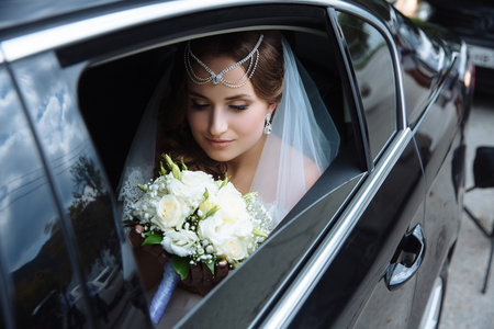 The bride is dressed in a wedding dress and veil, her head is draped with a lapel and sits in the car and admires the wedding bouquet.の写真素材