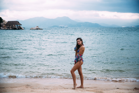 Beautiful young traveler posing on a white beach against a background of blue water in his new stylish bathing suit and beach shirt made from fine material. The girl enjoys her rest.の写真素材