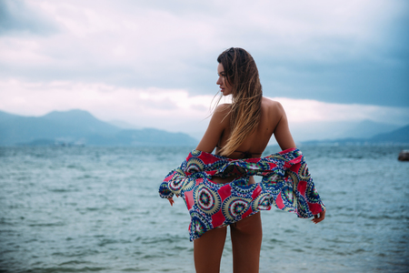 Rear view of a sexy slender model with long hair stands on the shores of the azure sea in a swimsuit and beach shirt, flattened from the shoulder. The fabric of the blouse neatly covers the girls ass, she smiles and enjoys the rest.の写真素材