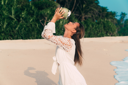 A close-up of coconut water flows over the face and neck of a young European girl with dark hair. A model on the beach pours coconut juice into his mouthの写真素材