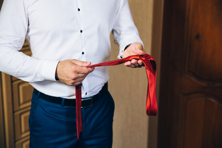 Close-up of a cropped frame of a slender business man in a trendy suit wearing dark trousers and a white shirt is holding a tie in his hands. A businessman is going to try on a red tie.の写真素材