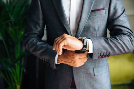 A close-up of a cropped frame of a stylish slender man in a jacket looks at his watch. Businessman is watching time, hurrying to an important meeting. Concept office, business, style, business suit.の写真素材