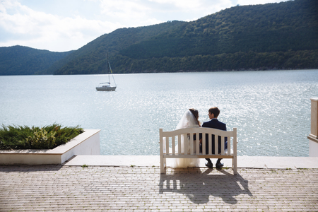 A view of the green hills and the blue calm river, along which the ship sails. On the beach on a white bench, the newlyweds admire the view, embrace and dream about the future.の写真素材