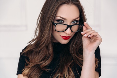 A close view of a young beautiful gray-eyed girl with scarlet lips holds her transparent glasses in black rim.の写真素材