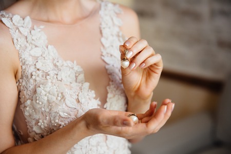Close-up of a cropped frame of a girl in a white lace dress with embroidery holding in hands a pearl earring. The bride is going to the weddingの写真素材