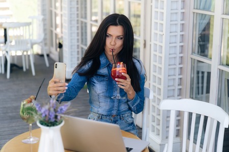 Front view of a bright brunette with long hair, has a European appearance, makes selfie on a smartphone, drinks her tasty cocktail through a straw, waits for her friends at a table in a cafe.の写真素材